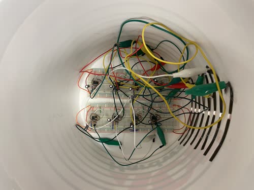 The breadboard embedded inside a plastic bucket which has holes cut in it and eight strips of reel-to-reel tape laced around it