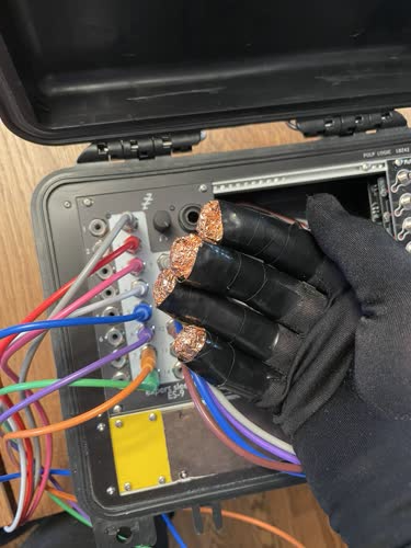 A glove with four fingertips replaced with conductive copper tape, which has been connected to modular synth cables that are going into the modular synthesizer in the background